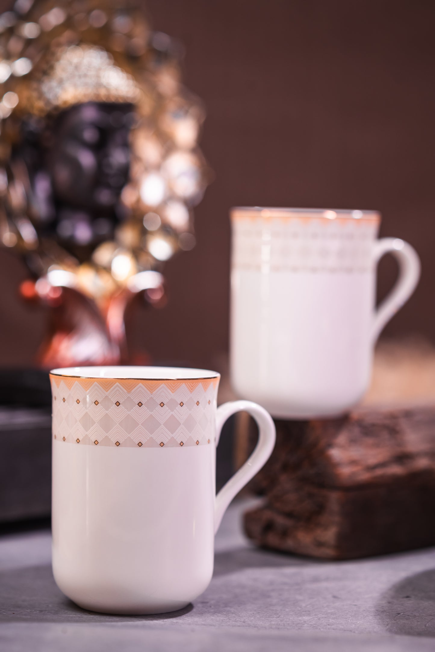 Two white tea/coffee mugs with decorative rims on a table with a blurred background