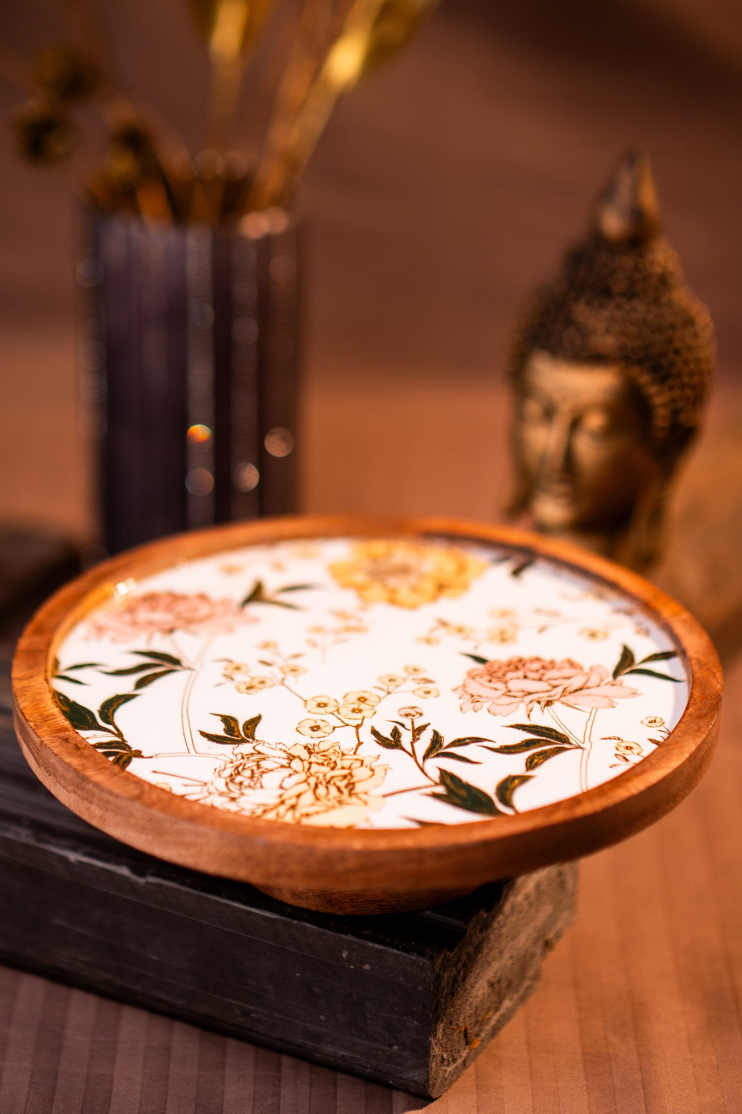 Wooden Cake Stand with floral design on a wooden stand, with a blurred Buddha statue in the background.
